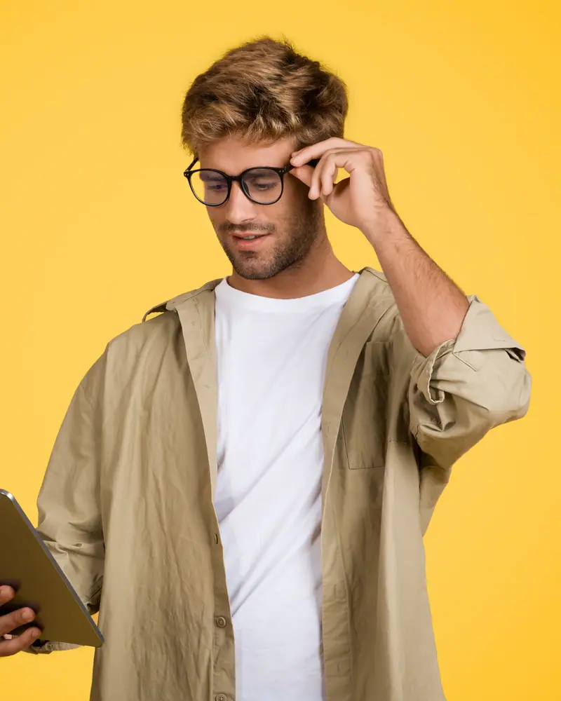 A man in glasses reading a tablet against a yellow wall