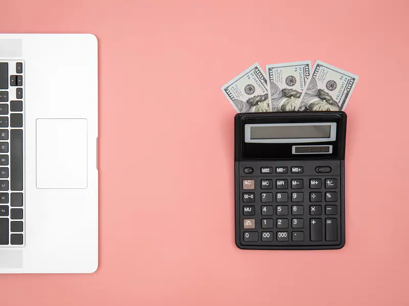 A calculator with banknotes next to a laptop on a pink desk